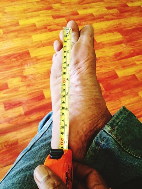 A person's bare foot being measured with a Brannock device, with a tape measure nearby, indicating careful at-home foot measurement.