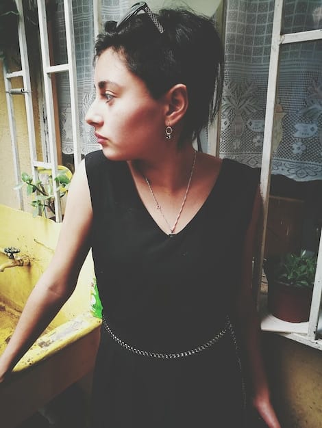 A woman with short, dark hair wearing a sleek black jumpsuit, adorned with oversized silver hoop earrings and a chunky chain necklace, at a rooftop bar at night.