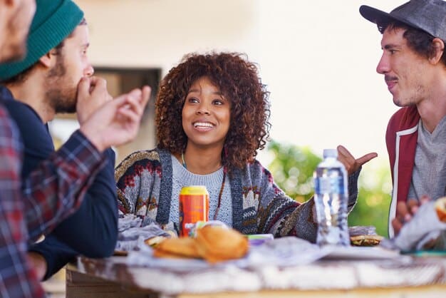 A diverse group of friends enjoying a casual outdoor brunch, with one person prominently featured in a stylish updated 2025 maxi dress, accessorized with a wide-brimmed hat and flat sandals, showcasing comfort and summer elegance.