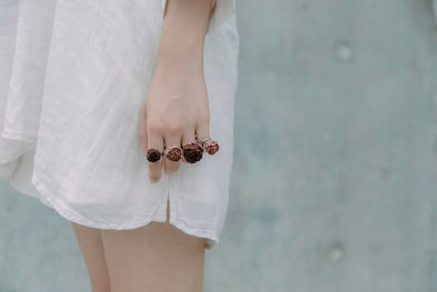 A close-up of a hand with various stackable rings on different fingers, including minimalist gold bands, one with a small pearl, and another with a delicate hammered texture, against a blurred, bright summer background.