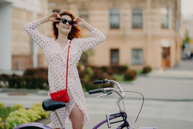 A young woman effortlessly riding a bicycle through a sunny park, wearing a light summer dress and a stylish, compact crossbody bag in a vibrant color.
